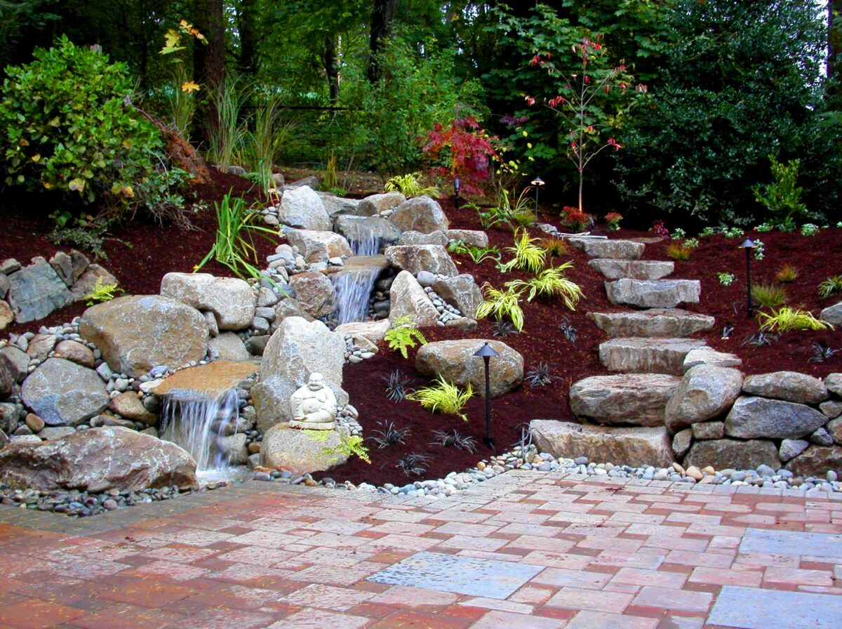 Custom waterfall fountain feature in residential backyard with large stone stairwell as seen landscaped with plants native to the Pacific Northwest and a paved patio.