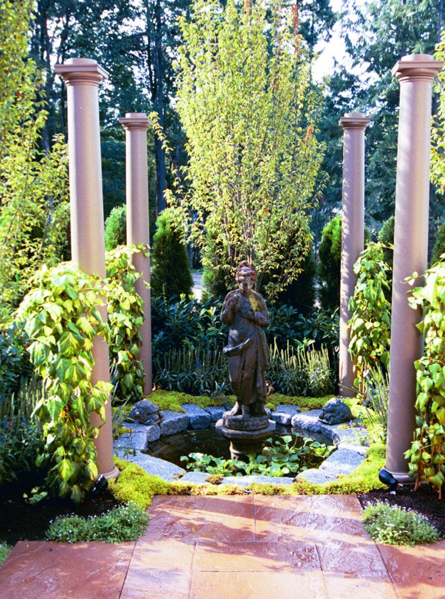 Circular lily pond with statue of a women in the center of the pond. The pond is lined with pillars and custom lighting to illuminate the scene. A paver patio lines the pond with lots of green landscaping.