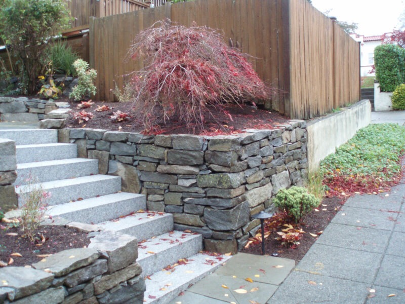 Rock retaining wall seen on either side of large granite stairwell leads to the front door of residential home in Seattle, Washington.