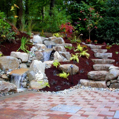 Custom waterfall fountain feature in residential backyard with large stone stairwell as seen landscaped with plants native to the Pacific Northwest and a paved patio.
