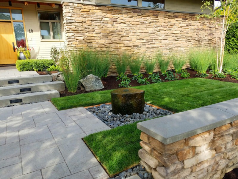 Small front yard filled with lush sod, unique rock feature water fountain, a paver patio leading up to the front door of a residential home in Seattle, WA.